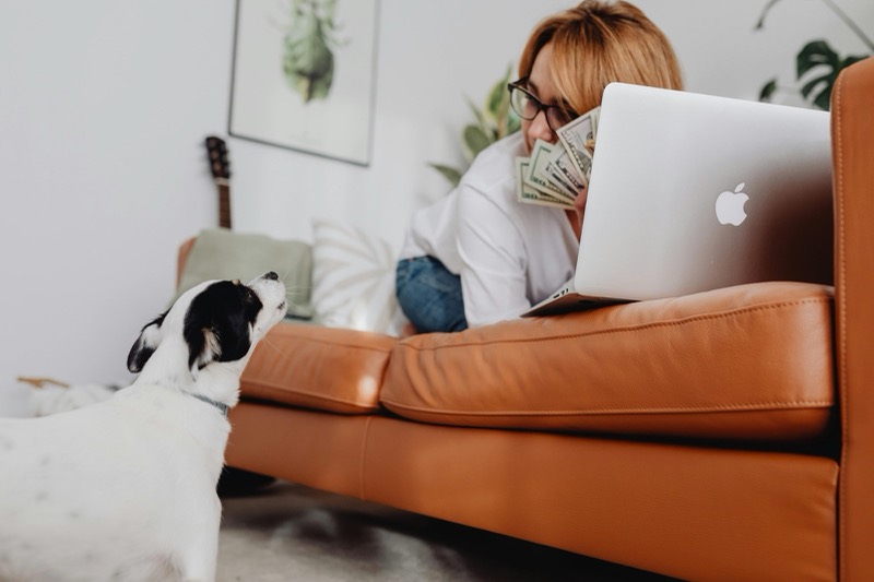 A woman on a sofa with a laptop and a fan of cash, watched by her dog