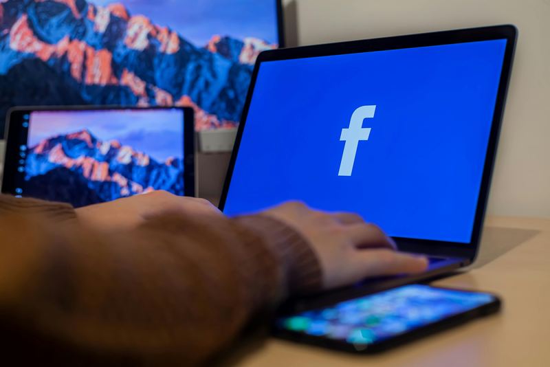A hand on a laptop showing the Facebook logo, with a phone and tablet beside it