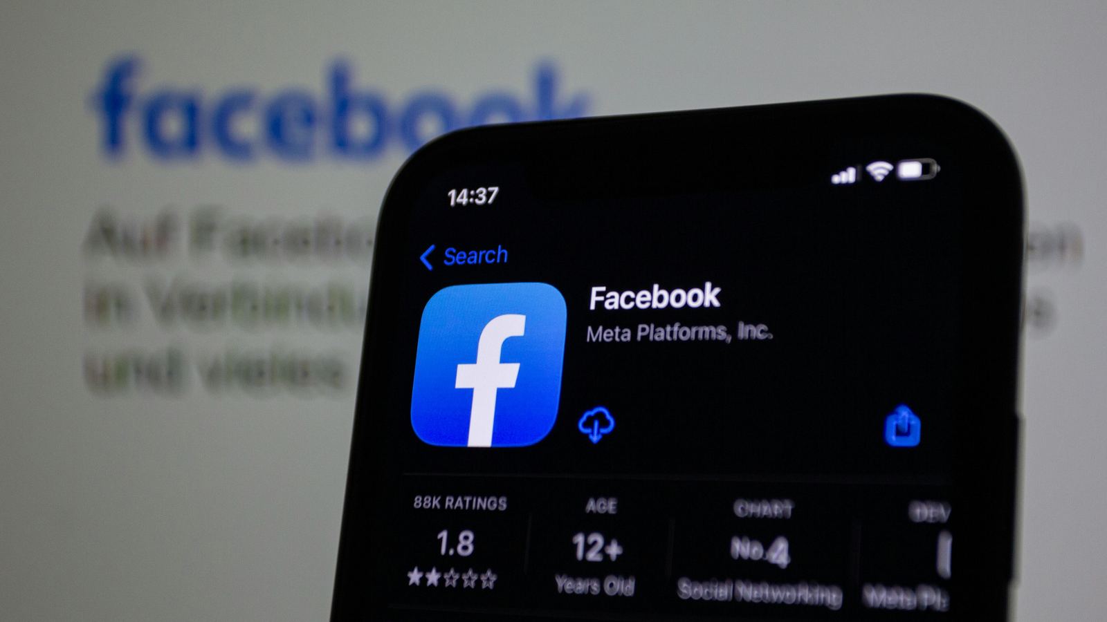 A phone showing the Facebook app page in front of a blurred Facebook logo