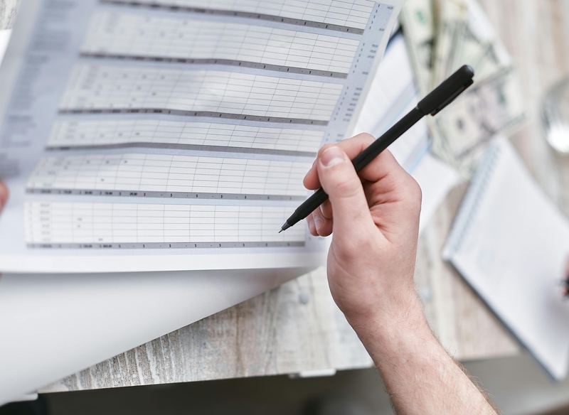 A hand holding a pen over a printed spreadsheet beside a notebook and cash