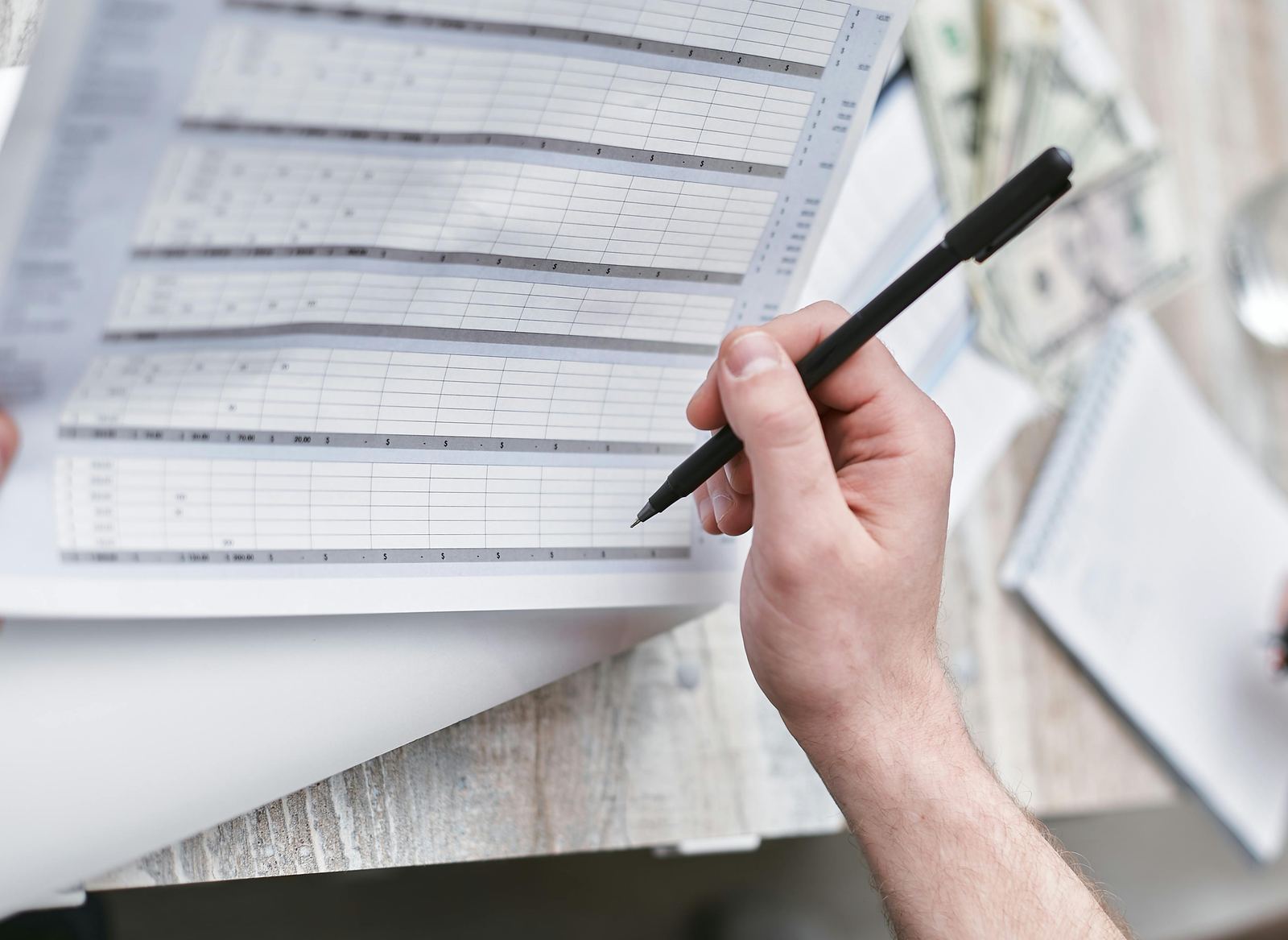 A hand holding a pen over a printed spreadsheet beside a notebook and cash